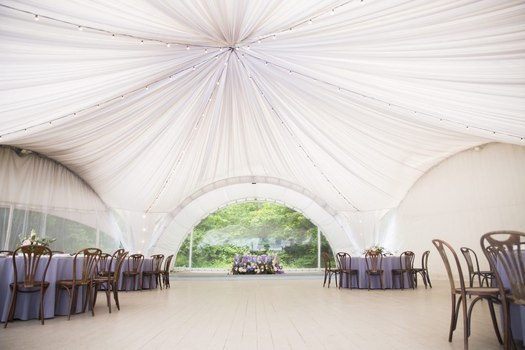 Big white wedding tent with beautiful decorations. Tables with floral decorations and wooden chairs